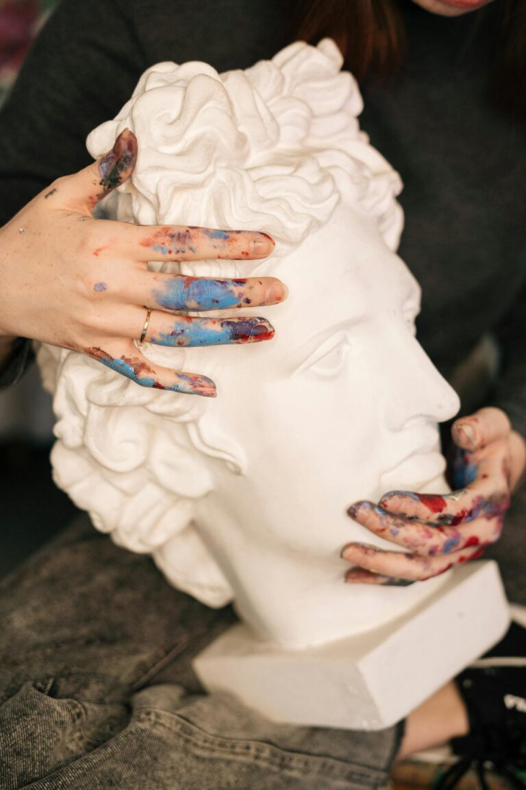 Hands with paint touching a classical sculpture head, showing creativity and artistry.